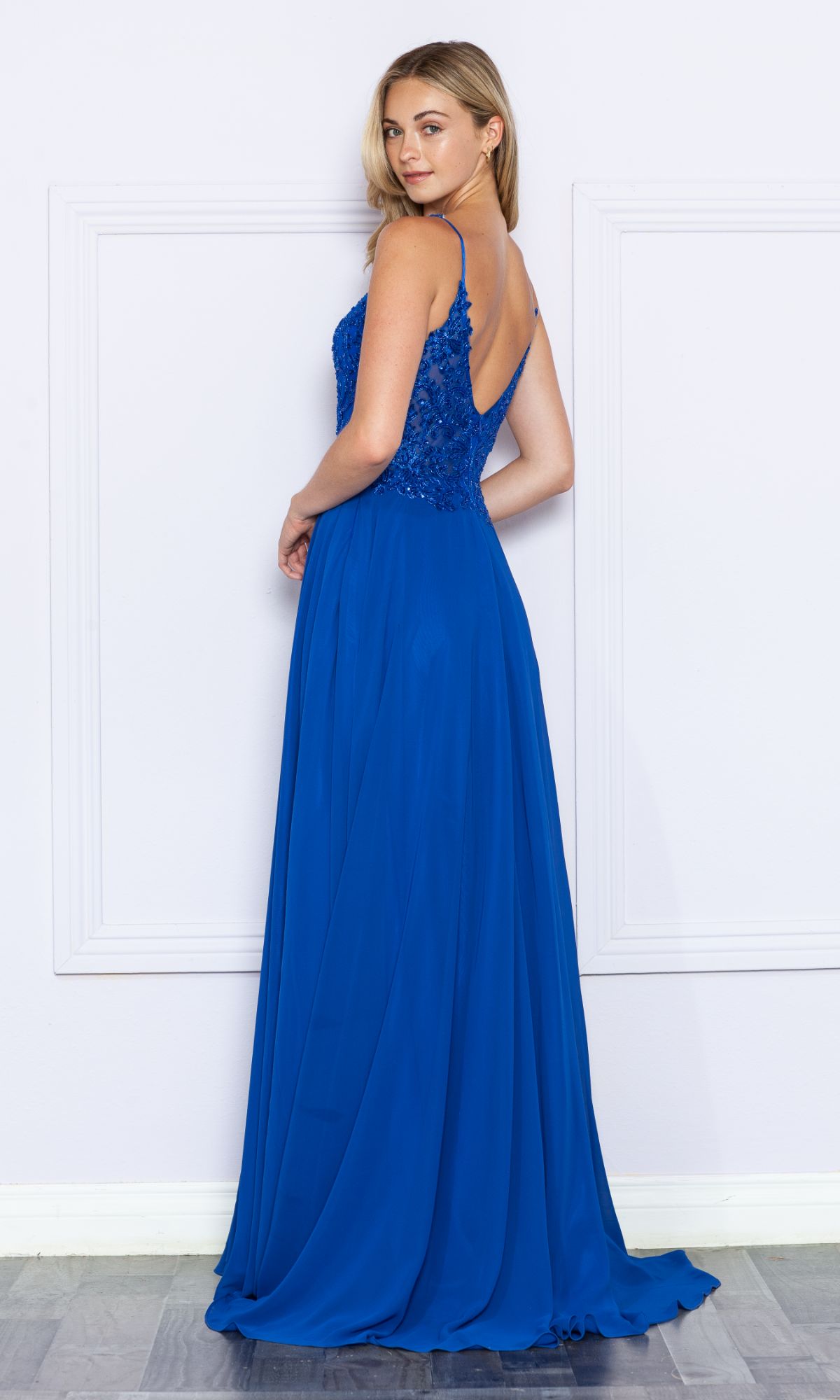 A woman poses against a pale wall wearing the Beaded-Bodice Long A-Line Prom Dress 9366, a sleeveless royal blue gown with a sparkly beaded bodice, lace detailing, floor-length skirt, and low back.